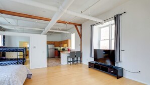 A spacious living area with a wooden floor, a large TV, and a kitchen in the background.