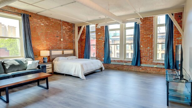 Une chambre à coucher moderne avec un mur en briques, un lit, un banc et un téléviseur posé sur une table en verre.