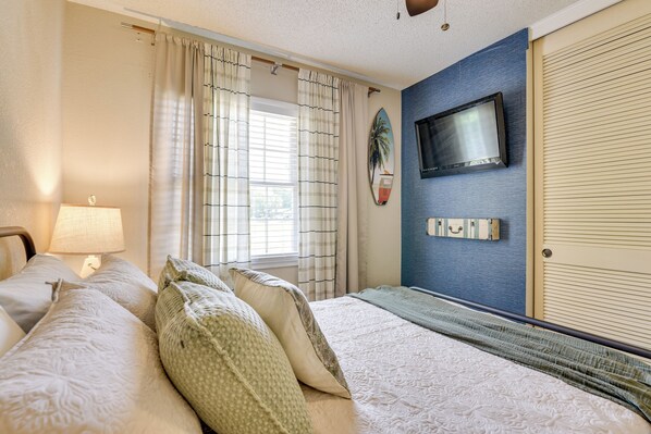 Habitación de hotel con cama, televisor montado en la pared, ventana con cortinas y lámpara en una mesita de noche.