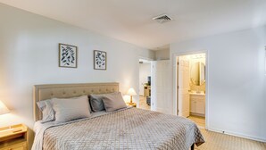Un dormitorio con cama, mesitas de noche, una puerta que da a un baño y una pared con dos cuadros enmarcados.