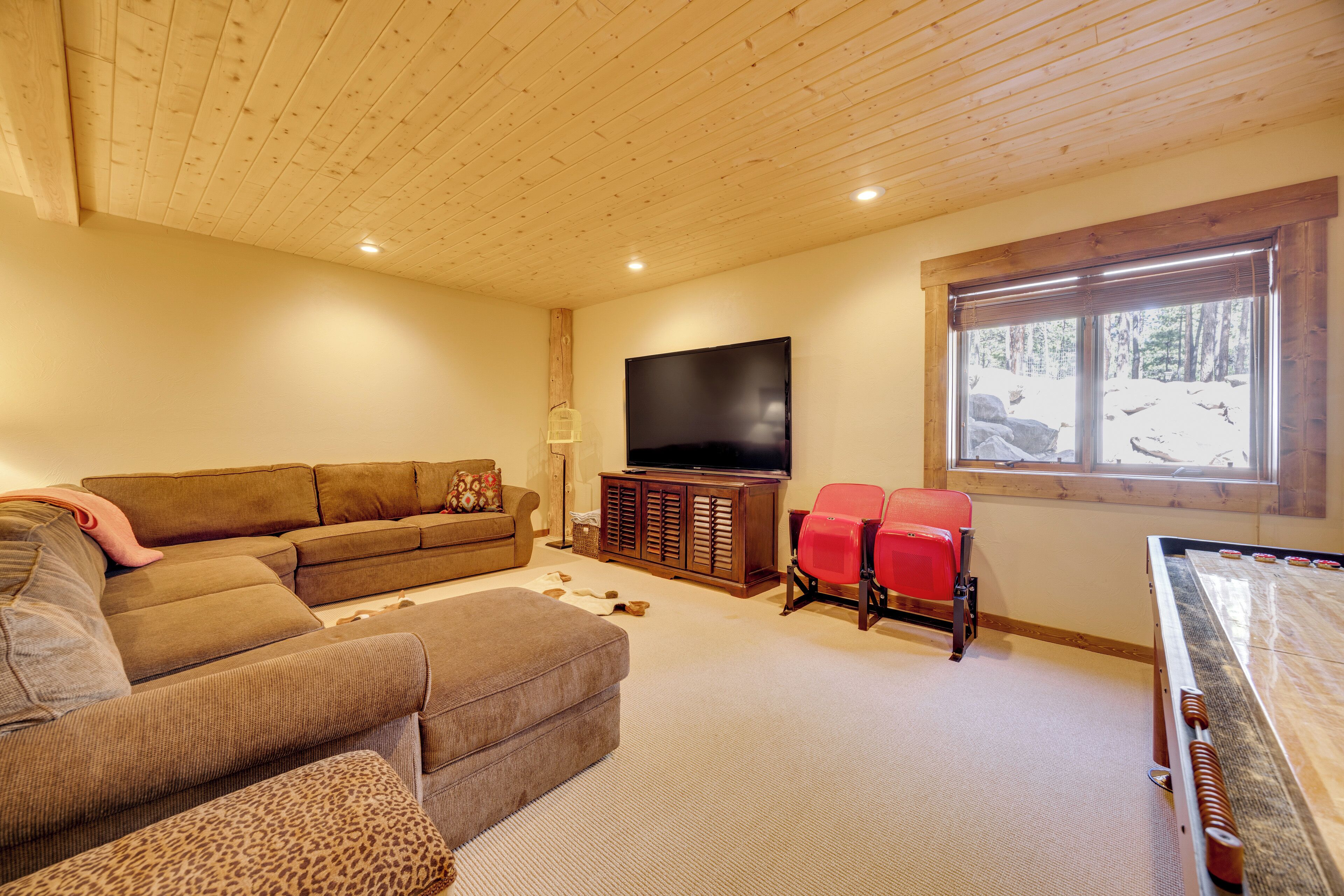 A cozy living room with a sofa, ottoman, and red chairs, a flat-screen TV, and a large window with a view of trees.