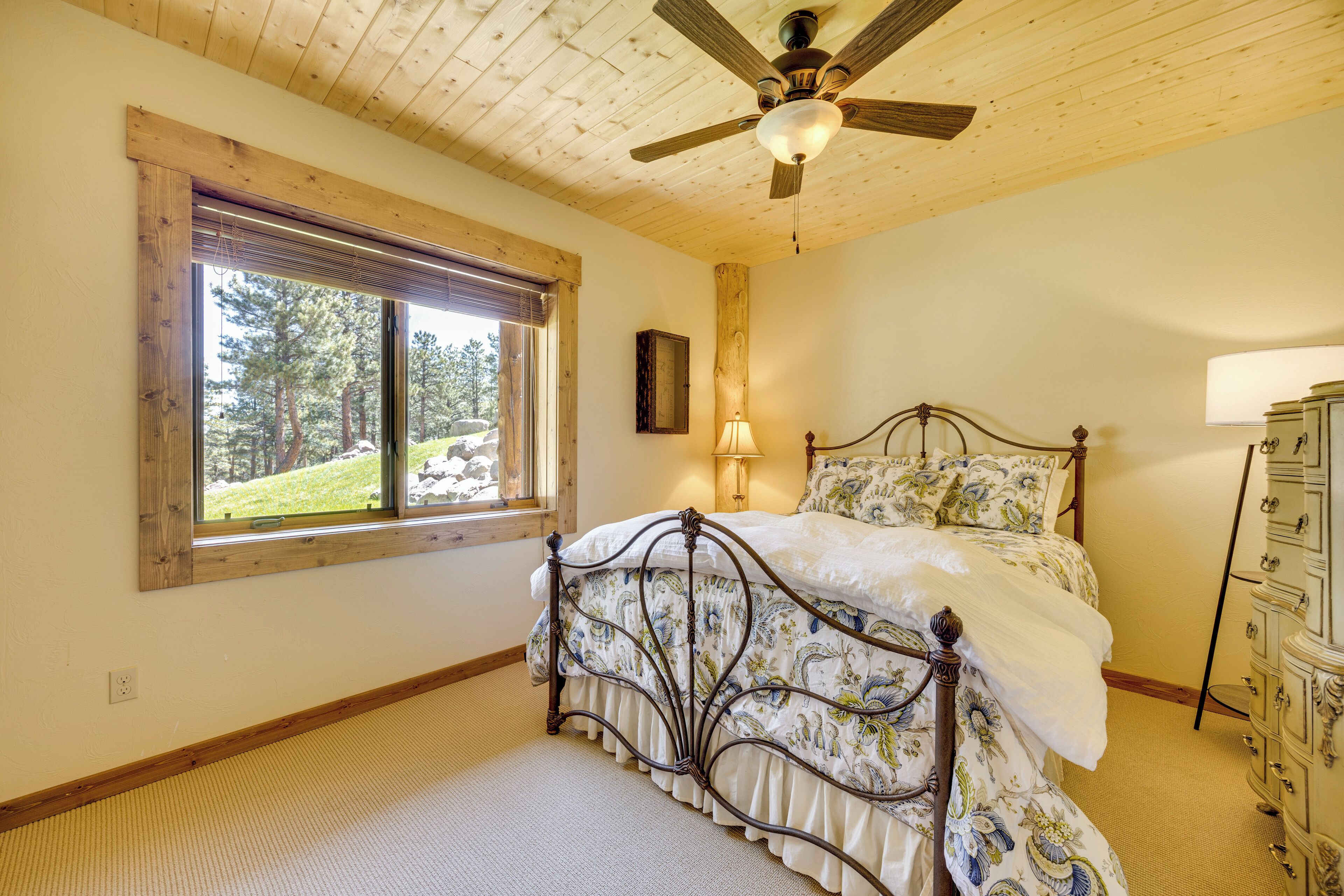 A bedroom with a wooden ceiling, a bed with floral bedding, a nightstand, a dresser, and a window with a view of trees.