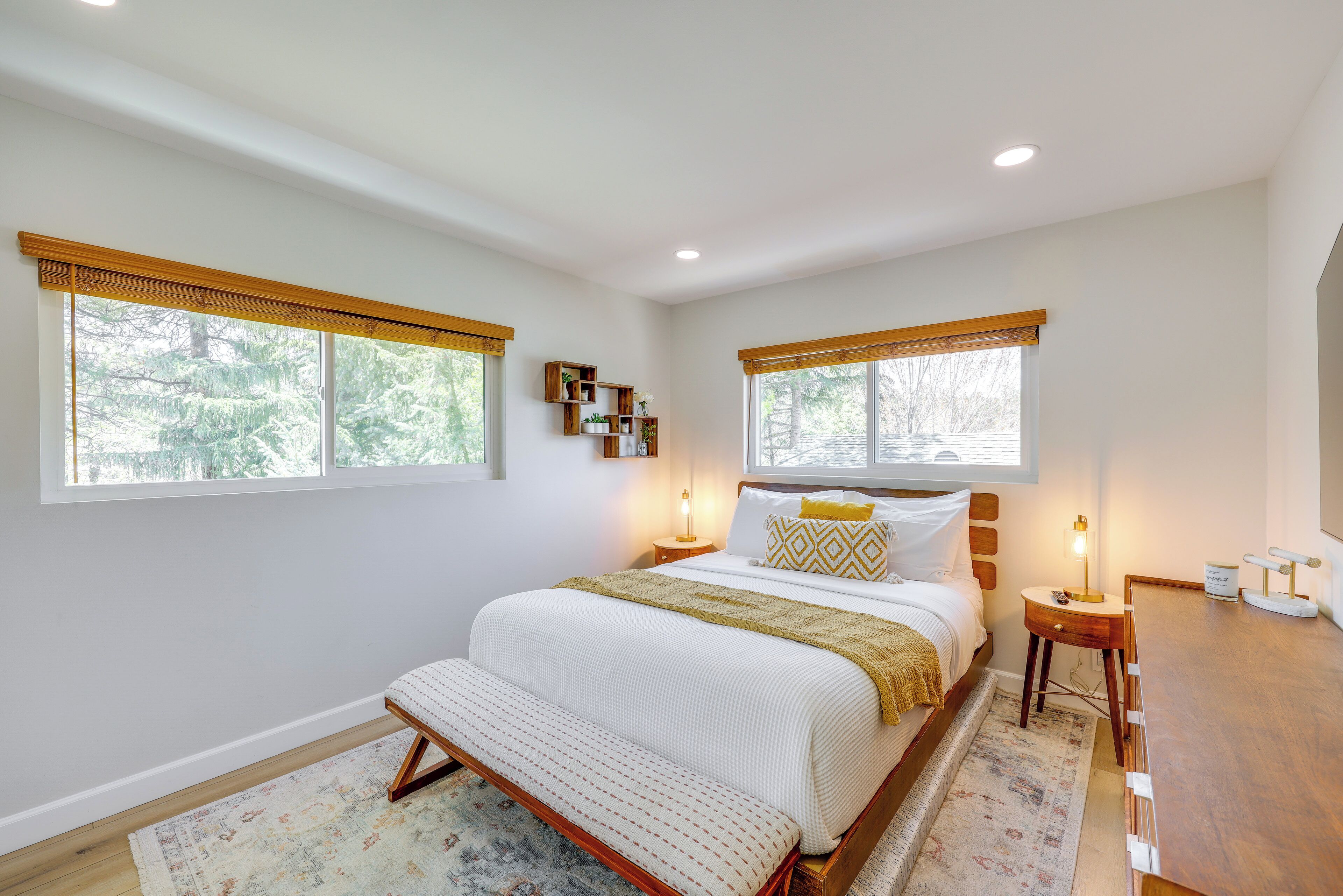 A bedroom with a large bed, two bedside tables, a nightstand, a wooden desk, and a window with a view of trees.