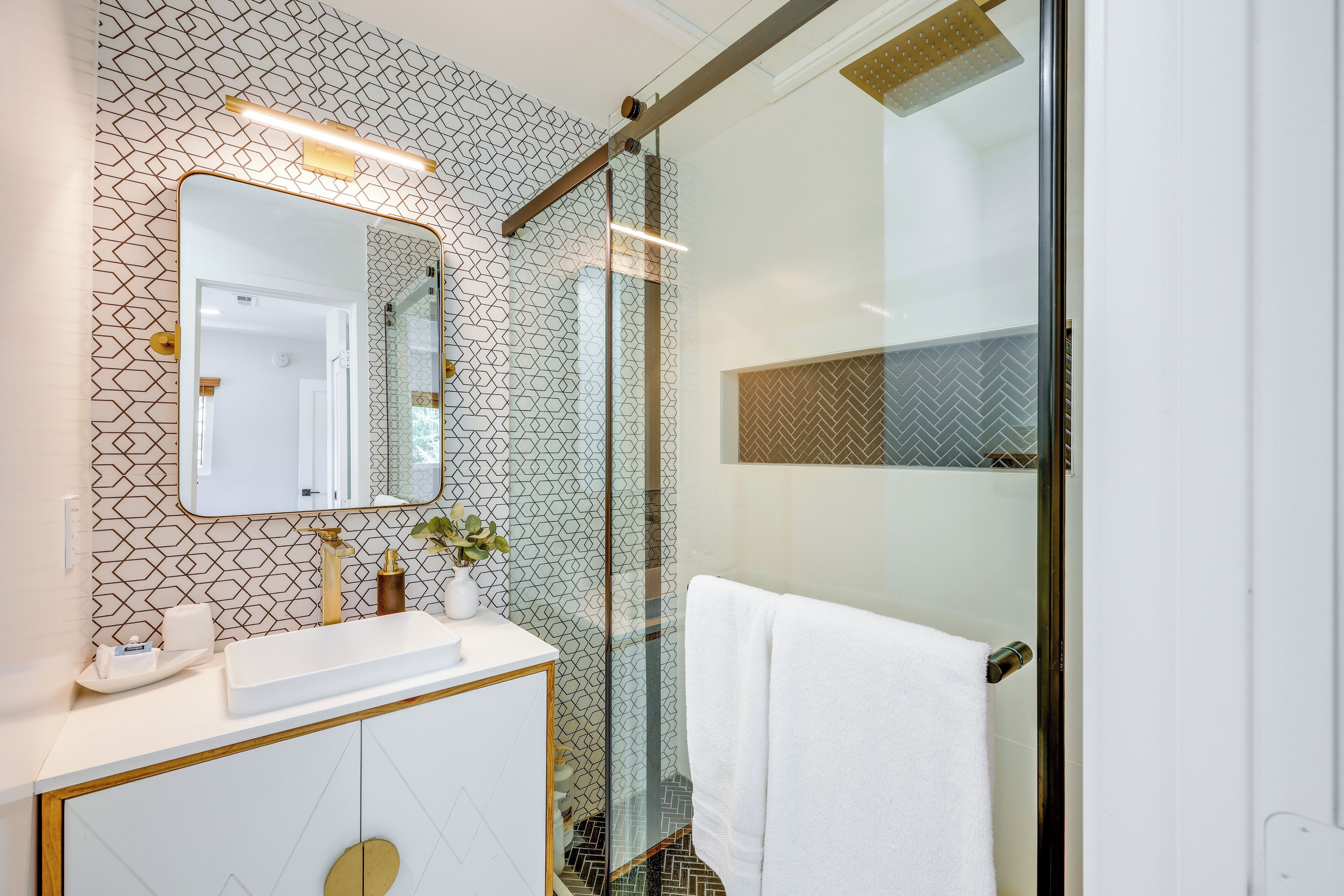 A modern bathroom with a geometric patterned wall, a large mirror, a white sink, and a glass shower enclosure.