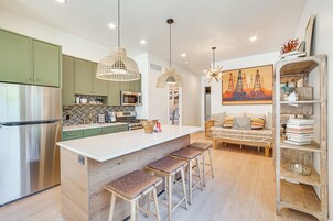 Cocina moderna con una isla central, armarios verdes, electrodomésticos de acero inoxidable y un comedor con sofá y taburetes de bar.