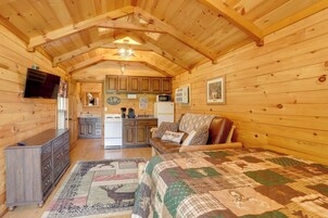 Interior acogedor de una cabaña de troncos con techo de madera, una cama con colcha, un aparador de madera, una zona de cocina y un televisor montado en la pared.