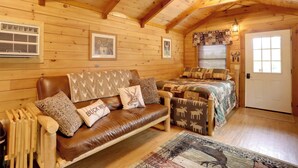 A cozy log cabin interior with a leather sofa, a bed with patterned bedding, wooden walls, and a ceiling with exposed beams. - 1 Mi to Current River: Cozy Studio Cabin! (Van Buren)