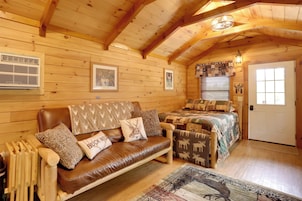 Un acogedor interior de cabaña de madera con un sofá de cuero, una cama con ropa de cama estampada, paredes de madera y un techo con vigas a la vista.