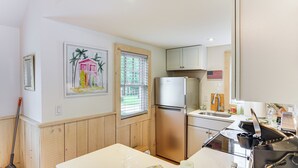 Eine Küche mit Kühlschrank, Spüle und Holzschränken. Ein Fenster mit Jalousien, ein Bild an der Wand und ein Besen, der an die Wand gelehnt ist.