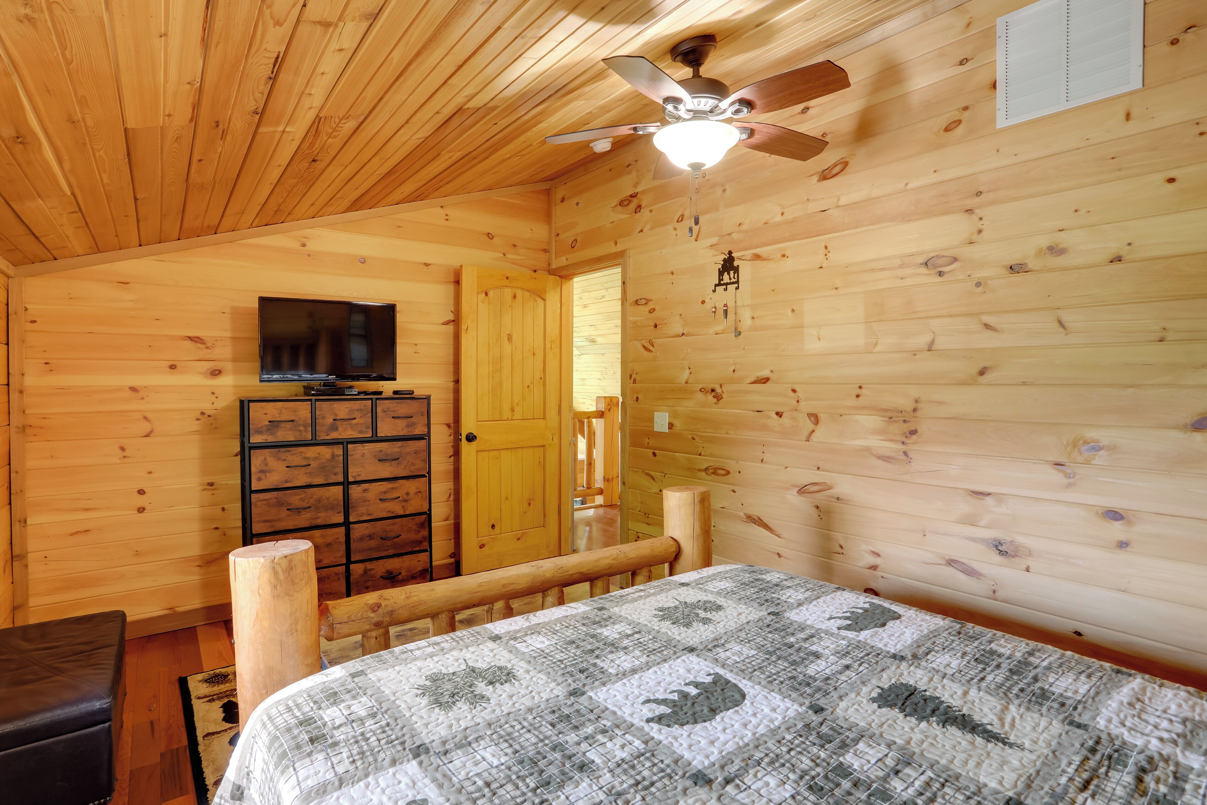 A wooden cabin room with a bed, a TV, a dresser, and a ceiling fan.