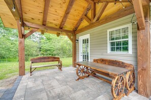 A covered patio with a wooden bench and a table with wagon wheel legs, set against a house with a white door and window.