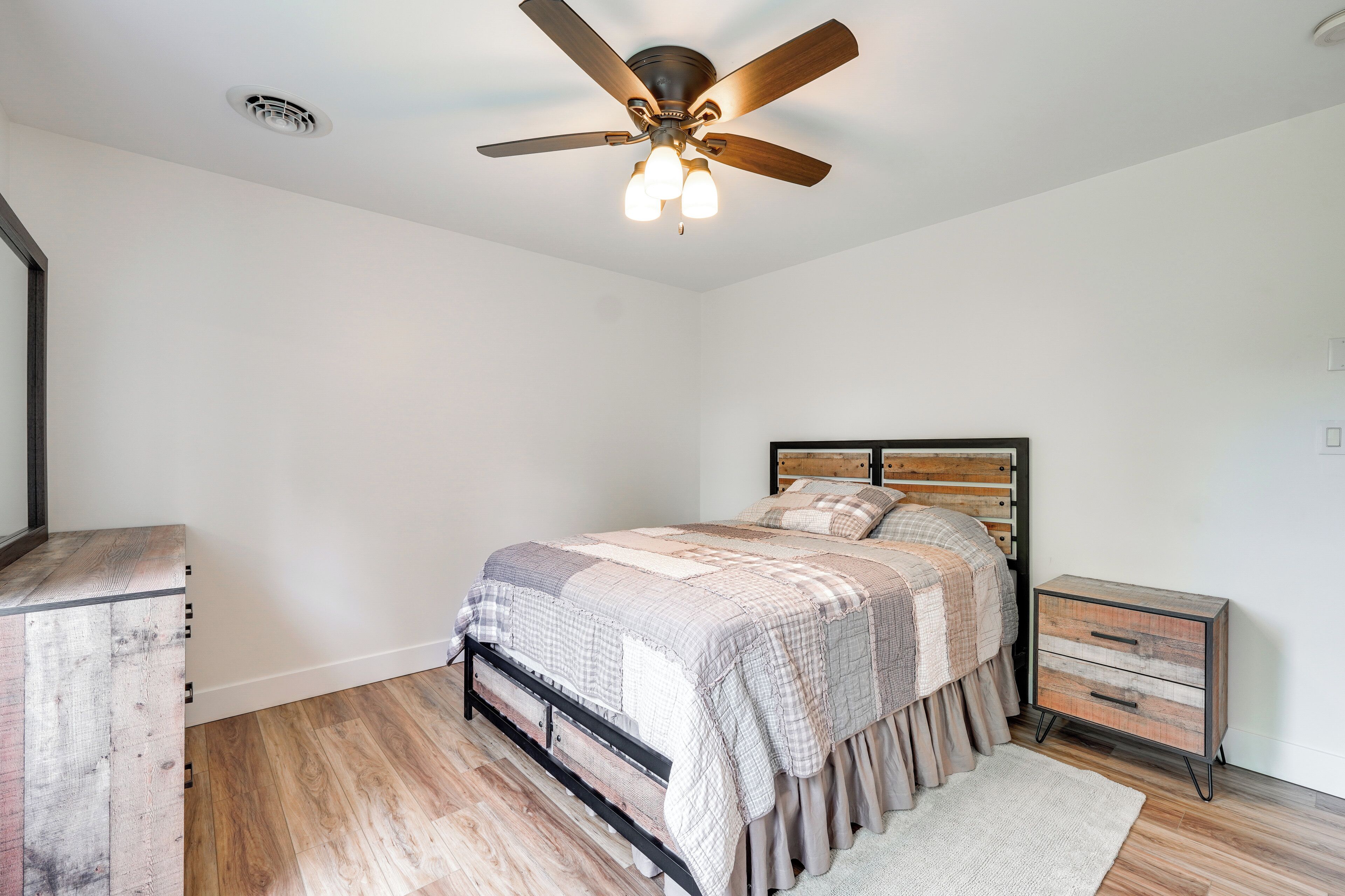 Un dormitorio con cama, mesita de noche, ventilador de techo y cómoda de madera.