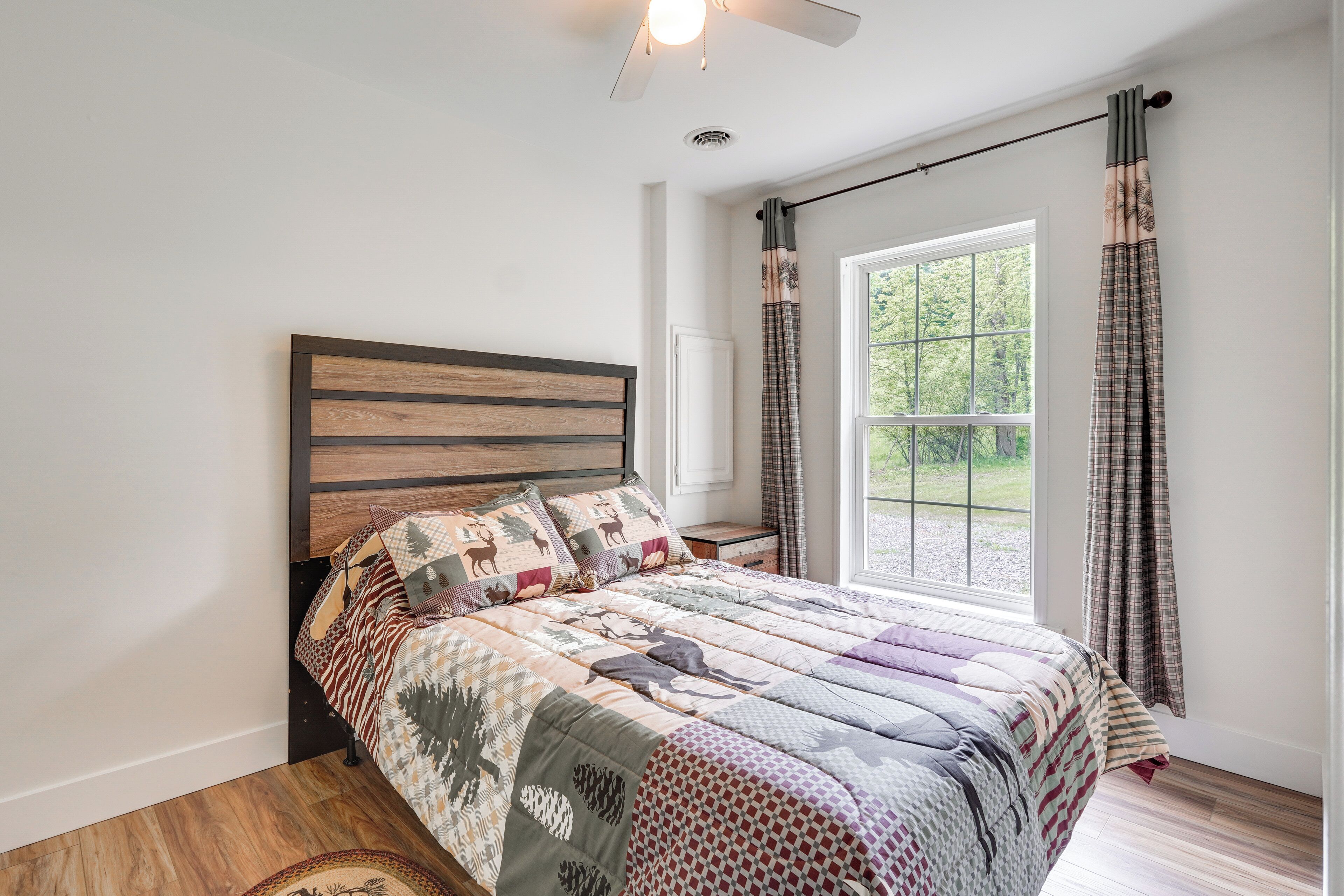 A bedroom with a wooden headboard, a large window with curtains, and a quilted bedspread.