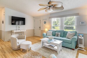 A living room with a teal sofa, white armchair, and ottoman, a white coffee table, and a ceiling fan. - 2 Blocks to Crystal Coast: Family-friendly Retreat (Harkers Island)