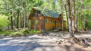 A cabin surrounded by tall trees, with a gravel driveway leading to the entrance.
