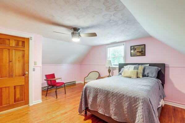 A bedroom with a wooden door, a red chair, a bed with a grey bedspread, a ceiling fan, and a window with a view of trees.