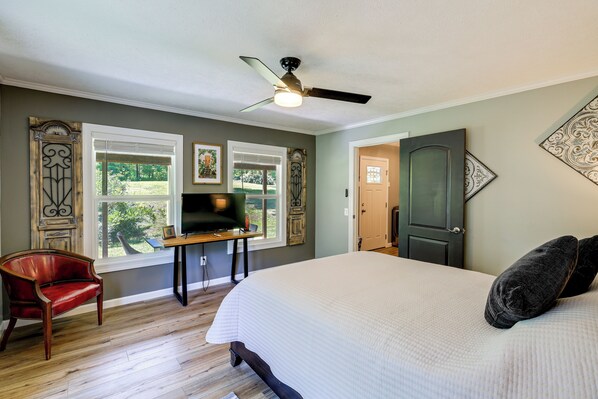 A bedroom with a large bed, a ceiling fan, a TV on a stand, a red chair, and a door leading to another room. - Creekside Retreat w/ Mtn Views in Lambsburg! (Lambsburg)