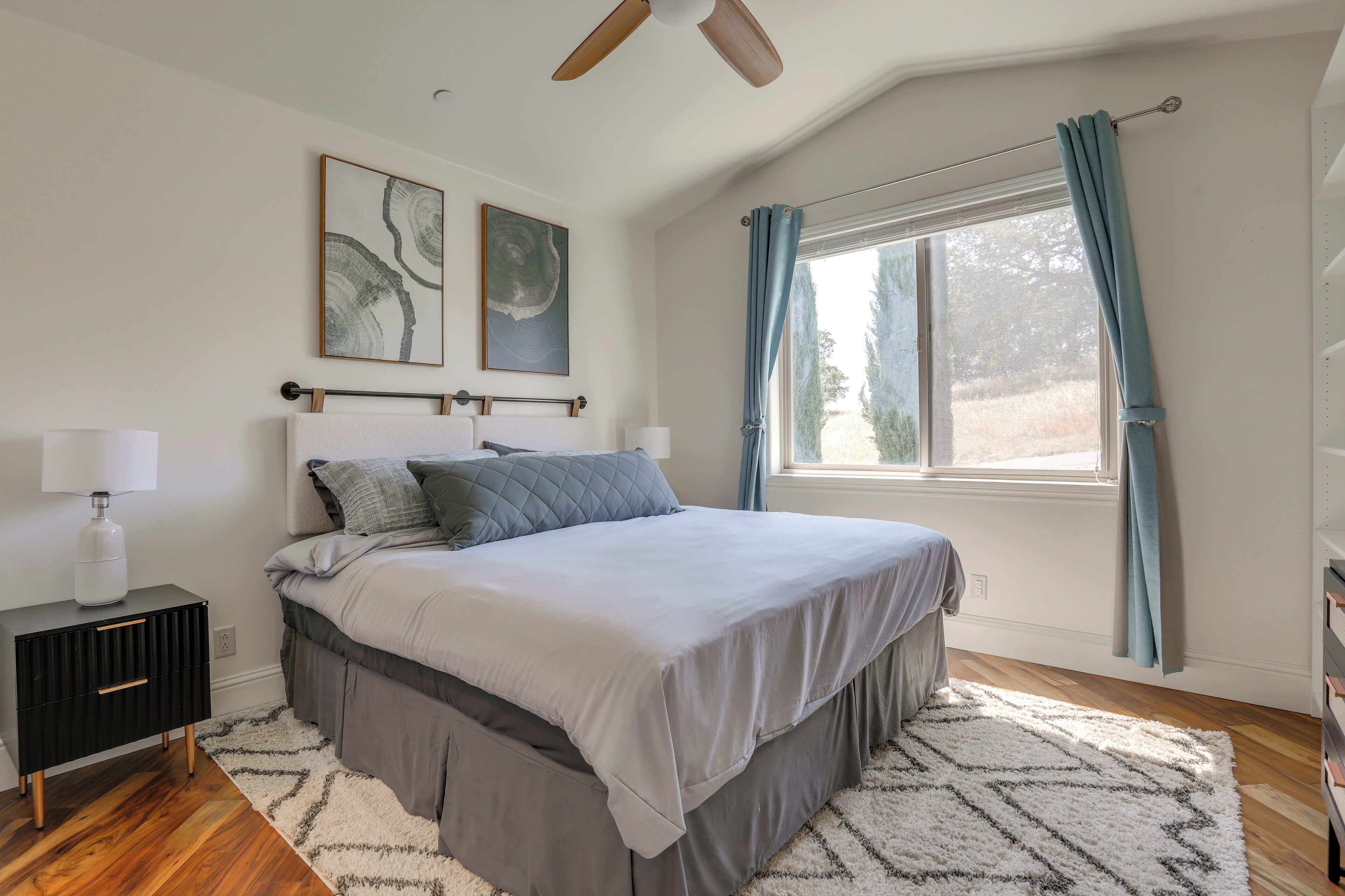 A bedroom with a large bed, a nightstand, a ceiling fan, and a window with curtains.