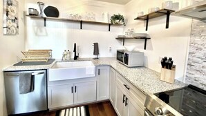 Una cocina moderna con armarios blancos, encimera de granito, refrigerador de acero inoxidable, microondas y horno. Hay estantes sobre la encimera con vajilla y una planta en maceta.