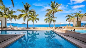 Área de piscina com espreguiçadeiras e palmeiras, com vista para um corpo de água.