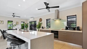 A modern kitchen with a central island, bar stools, ceiling fans, and a large window with a view. - Modere - Pool Entertainer Child Pet (Cairns)
