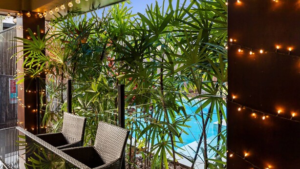 A balcony with wicker chairs, string lights, and a view of a swimming pool surrounded by lush green plants. - Modere - Pool Entertainer Child Pet (Cairns)