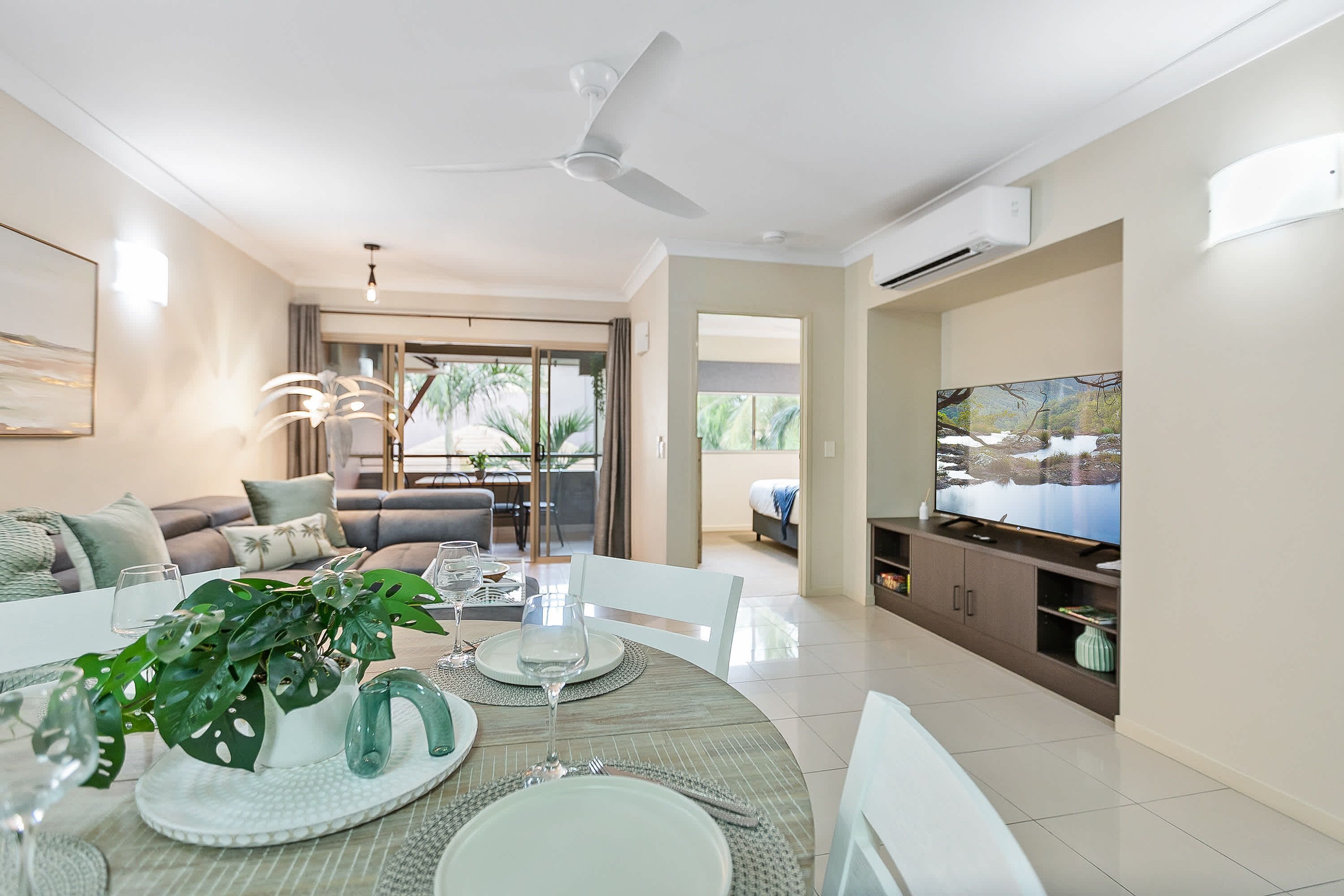 A modern living room with a dining area, a sofa set, a TV, and a balcony with a view of greenery.