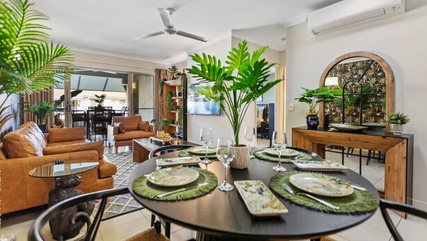 Un coin repas avec une table ronde dressée pour quatre personnes, entourée de chaises. Des plantes en pot sont posées sur la table et un ventilateur de plafond est installé au-dessus.