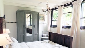 A bedroom with a bed, a nightstand, a lamp, a wardrobe, a chair, and a window with curtains. - L'Ecurie - The original farmer's cottage (Larzac)