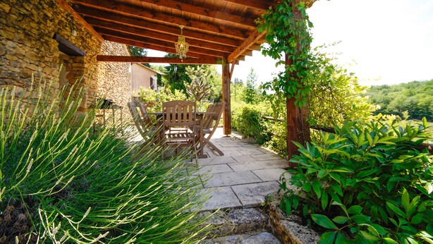 A covered patio with stone flooring, wooden furniture, and lush greenery. - L'Ecurie - The original farmer's cottage (Larzac)