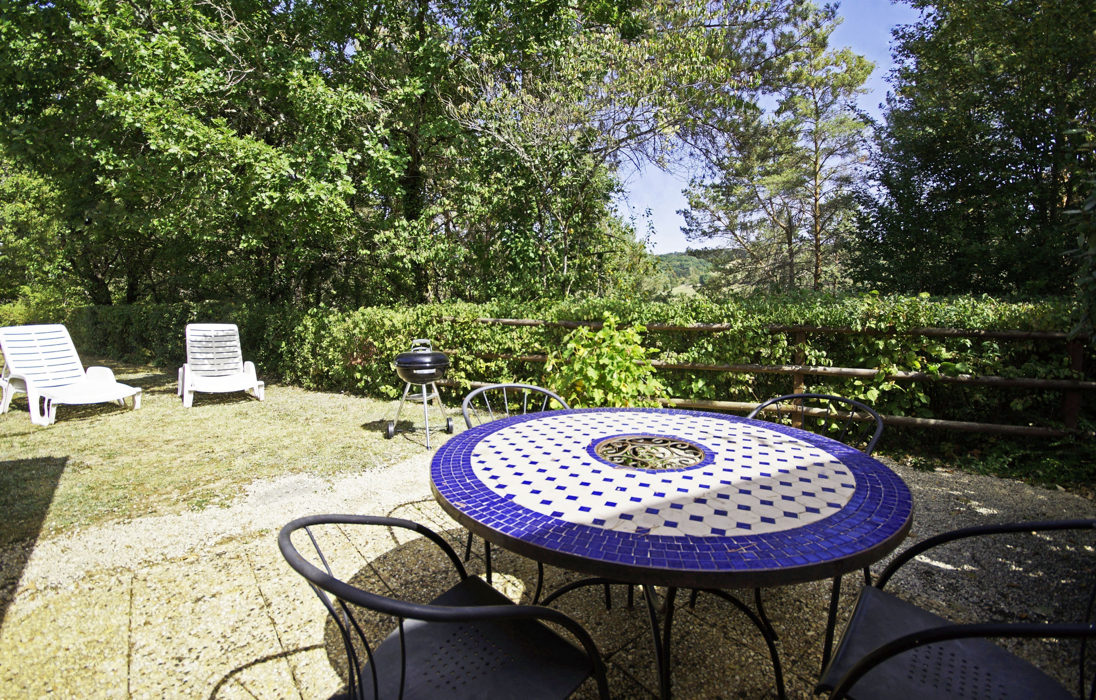 An outdoor seating area with a round table and chairs, surrounded by greenery and a wooden fence.