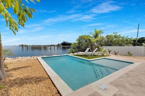 A rectangular swimming pool with a clear blue water surface, surrounded by a pebble beach and a wooden deck.