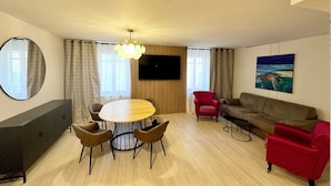 A modern living room with a round wooden table, four chairs, a red armchair, a brown sofa, a mounted TV, and a painting on the wall.