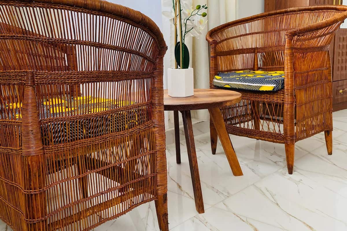 Mobiliario de mimbre con un florero, un cojín estampado y una mesa de madera en una habitación con suelo de mármol blanco y ventanas grandes.