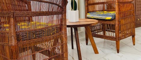 Mobiliario de mimbre con un florero, un cojín estampado y una mesa de madera en una habitación con suelo de mármol blanco y ventanas grandes.