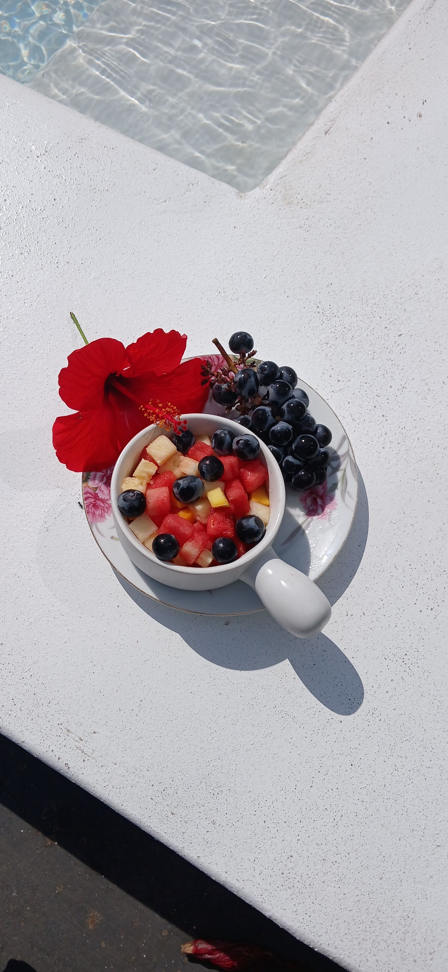 A bowl of fruit on a white table by a pool.
