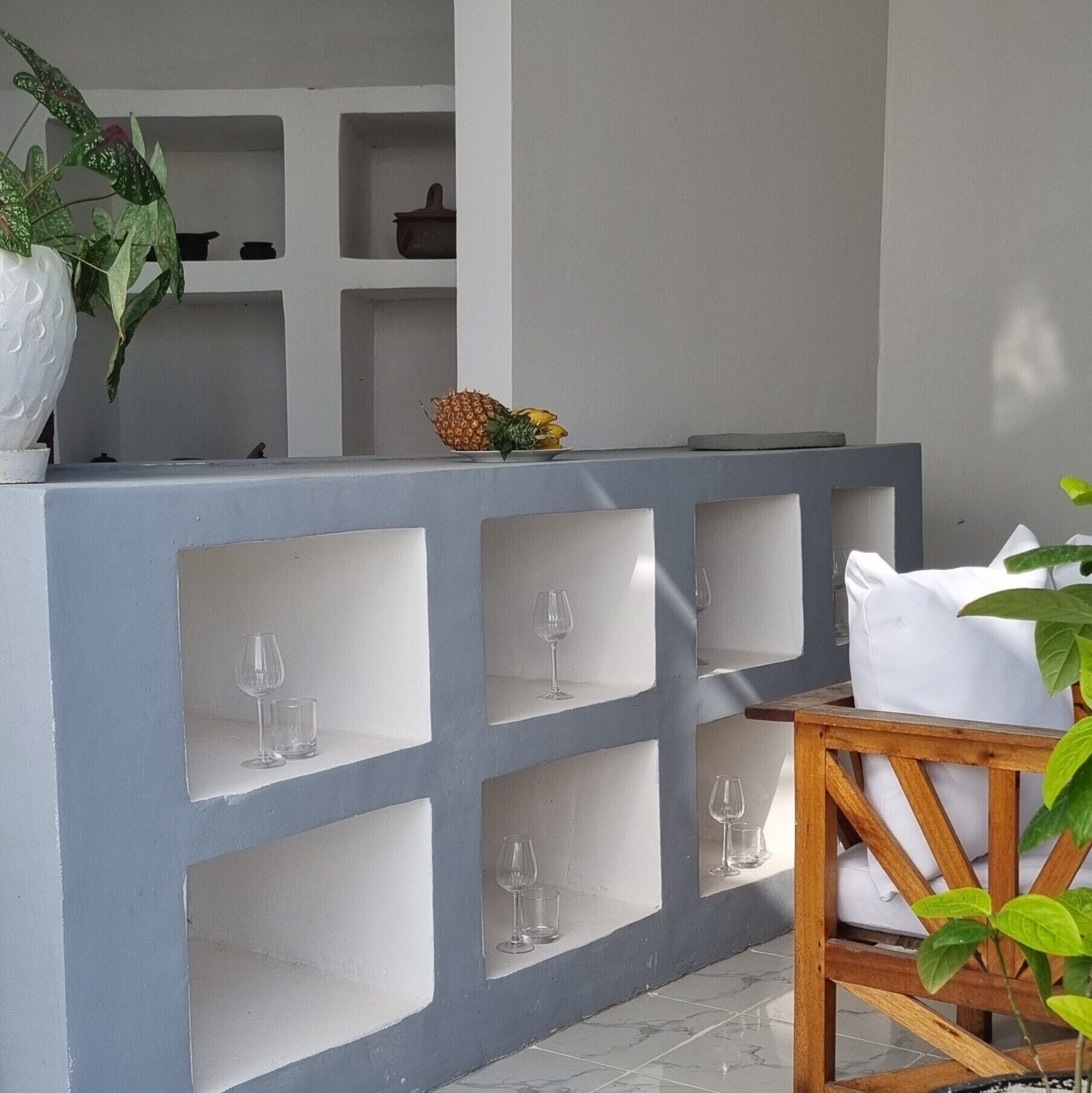 A modern outdoor seating area with a white and grey color scheme, featuring a wooden table and chairs, and multiple wine glasses displayed in built-in shelves.
