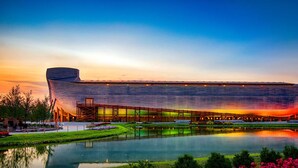 Un grand bâtiment moderne doté d’un toit incurvé original, situé à côté d’une étendue d’eau aux reflets changeants, entouré de verdure et surplombé par un ciel dégagé.