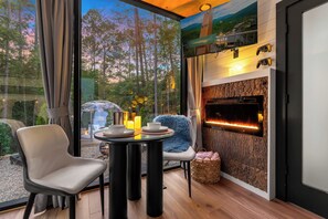 Un intérieur moderne avec une cheminée, une table à manger avec des chaises et une vue sur une forêt, visible à travers de grandes fenêtres.