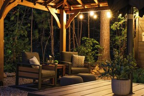 Terraza de madera al aire libre con dos sillas y una mesa, rodeada de árboles y una estructura de madera.