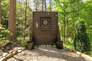 A wooden structure with a dartboard, surrounded by rocks and potted plants in a forest setting. - Villa Sol-Mirror Cabin-Private-Hot Tub-FirePit-Luxury (Sevierville)