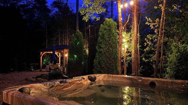 A hot tub in a forested area with a gazebo and string lights.