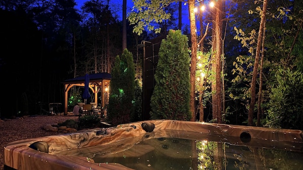 A hot tub in a forested area with a gazebo and string lights.