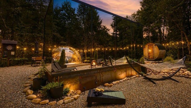 Entorno rústico al aire libre con una estructura espejada que refleja un espacio de estar acogedor, una hamaca y un barril de madera.