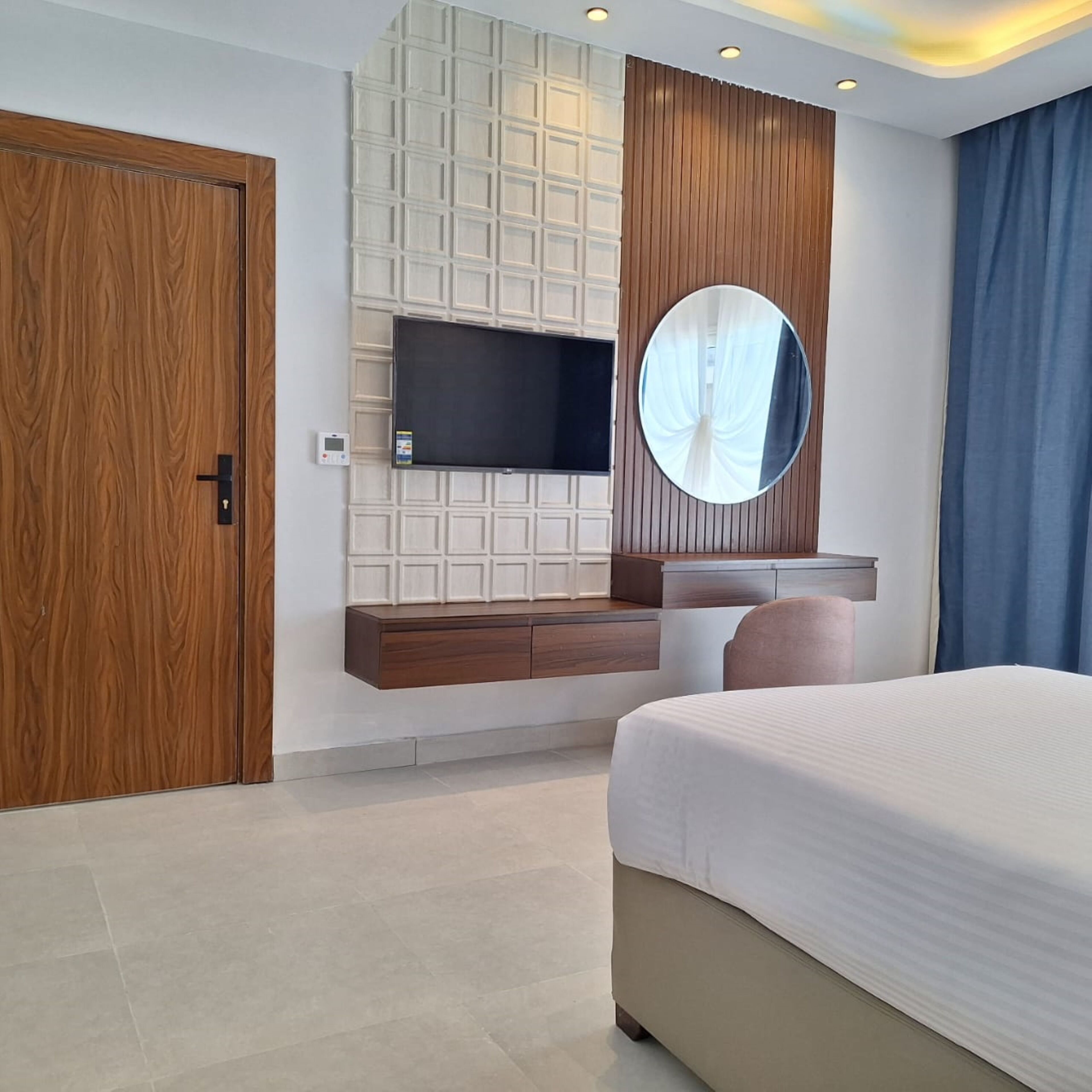 A modern bedroom with a large bed, a TV mounted on a wooden panel wall, a round mirror, and a built-in vanity with a sink.