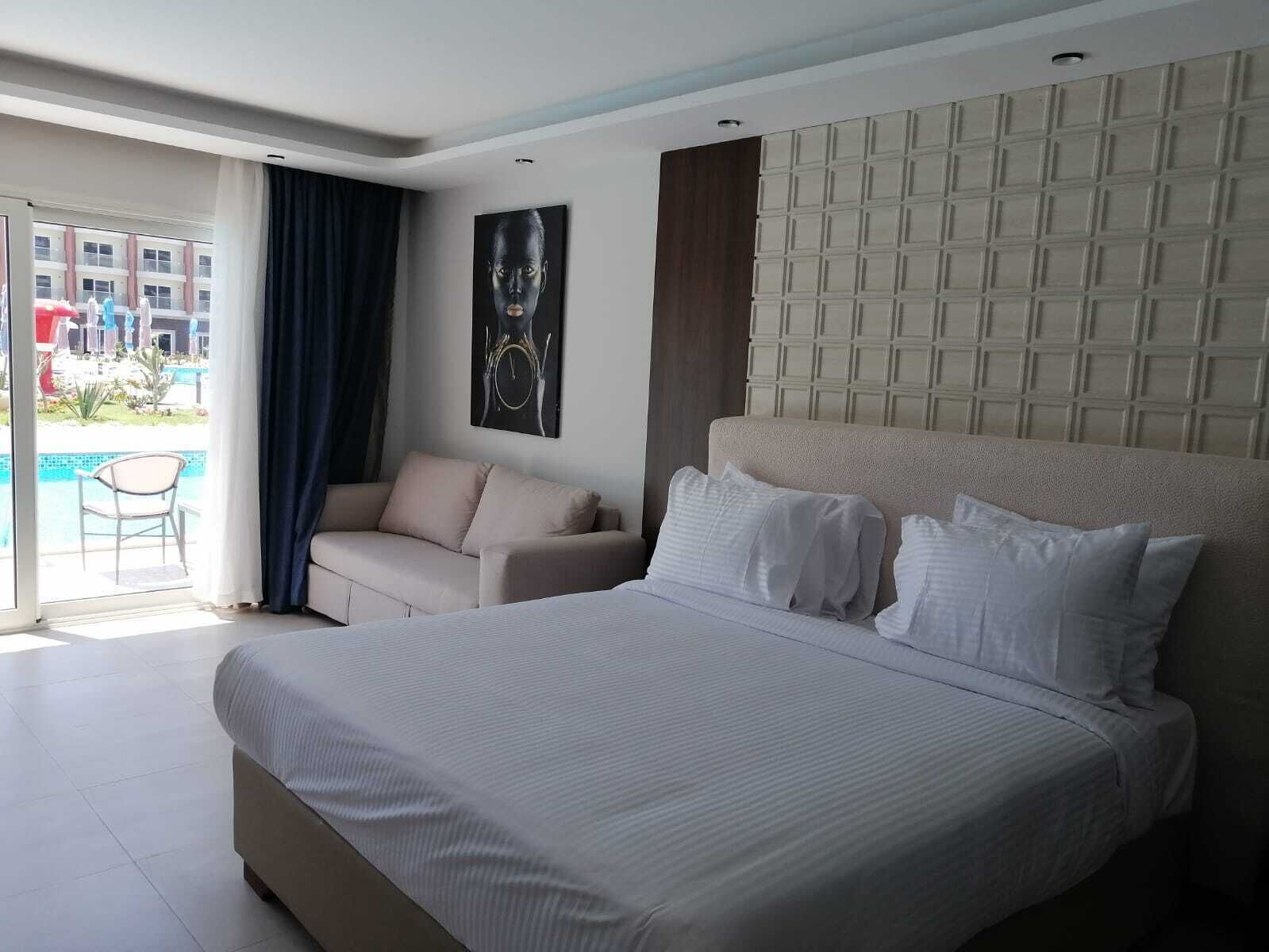 Habitación de hotel con una cama grande, un sofá, vista a una piscina y un paisaje urbano al fondo.