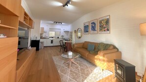 Ein modernes Wohnzimmer mit einer Couch, einem kleinen Tisch und einem Kamin. Im Hintergrund ist eine Küche mit einer Mikrowelle und einem Spülbecken zu sehen.