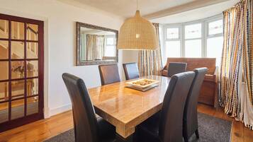 A dining area with a wooden table, four chairs, a large pendant light, a mirror, and a window with curtains.