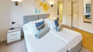 A neatly made bed with white linens and decorative pillows, a bedside table with a lamp, and a wall-mounted light fixture.