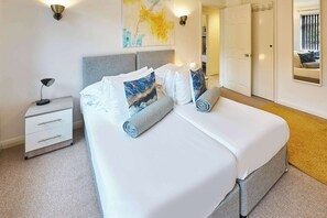 Un lit bien fait, avec du linge de lit blanc et des coussins décoratifs, une table de chevet avec une lampe et un luminaire fixé au mur.